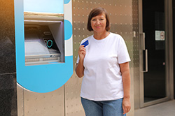 Beautiful middle aged woman smiling showing credit card near ATM outdoors